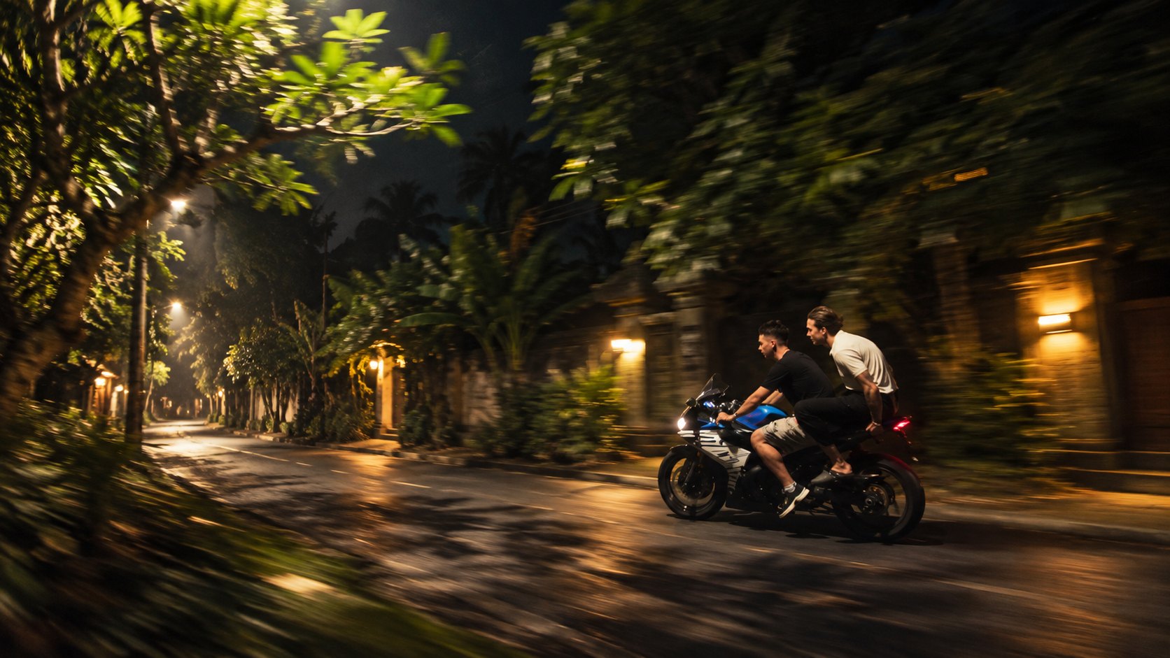 Two riders on a motorcycle moving quickly along a tropical residential road at night in Bali.