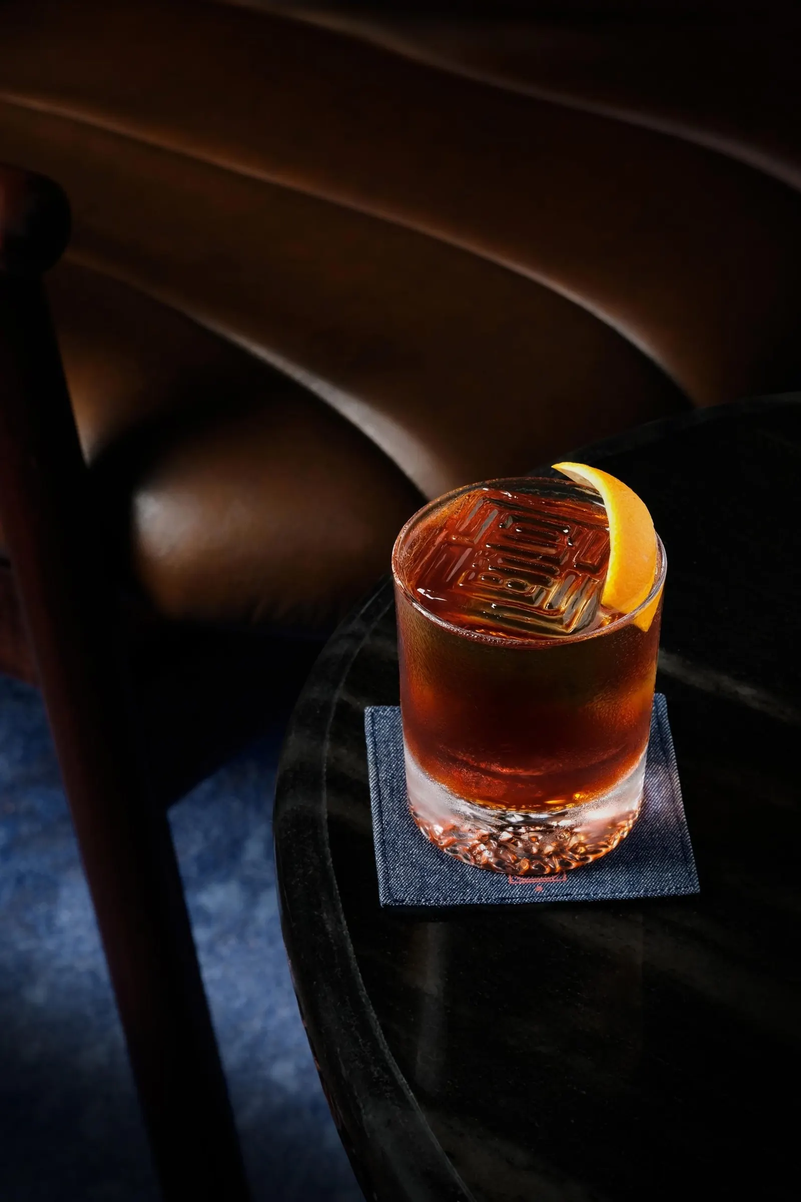 A stirred cocktail with orange on an AOKU coaster beside a booth.