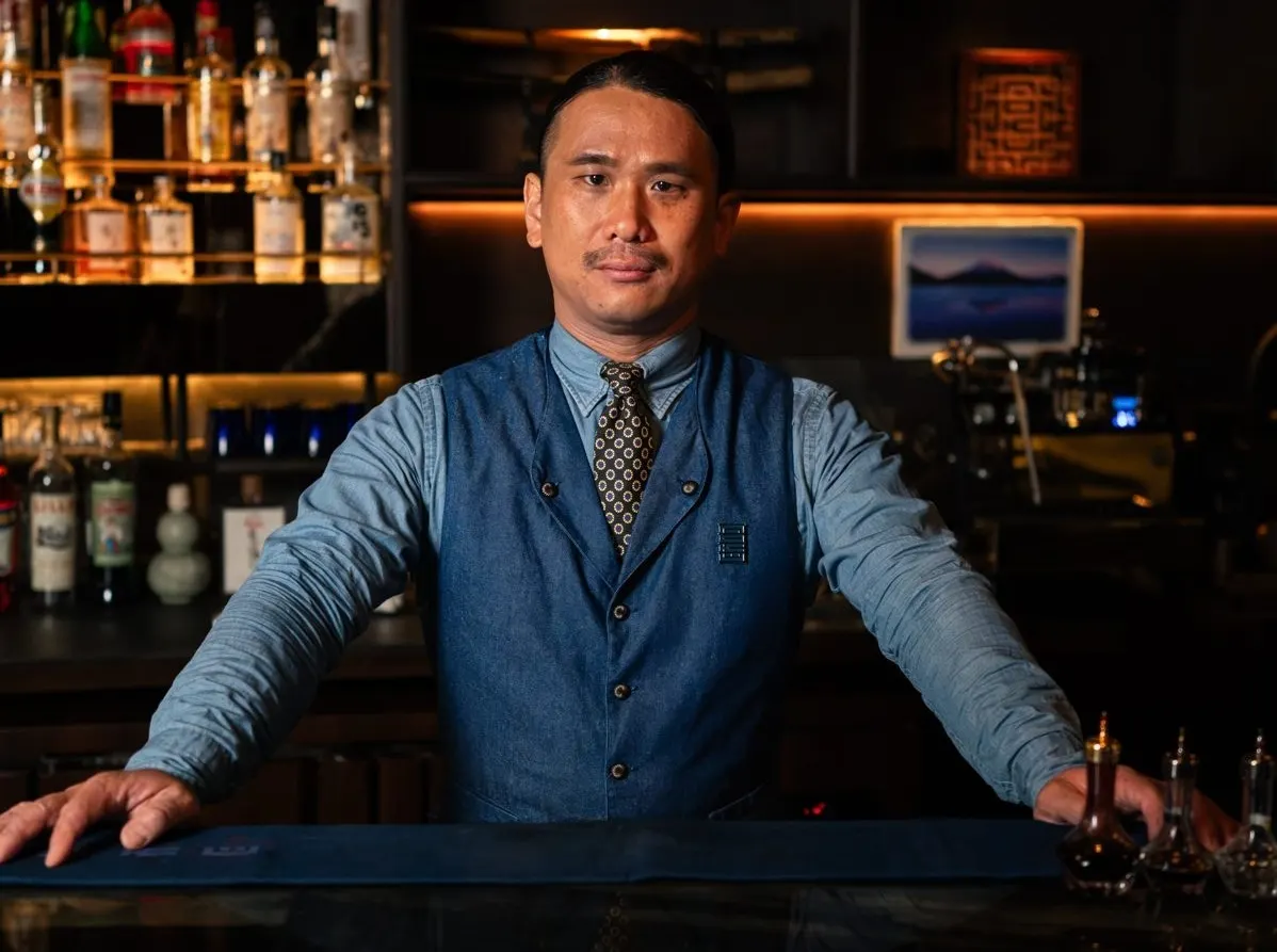 Kensho Hirai in a denim waistcoat and tie, leaning on the bar at AOKU.